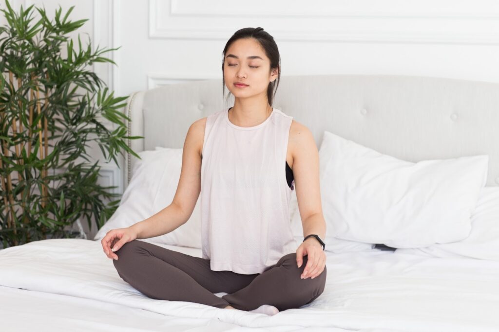 Woman sitting cross-legged on a bed with eyes closed, meditating in a bright room with white bedding and green plants, creating a calm and peaceful atmosphere: PMR Relaxation