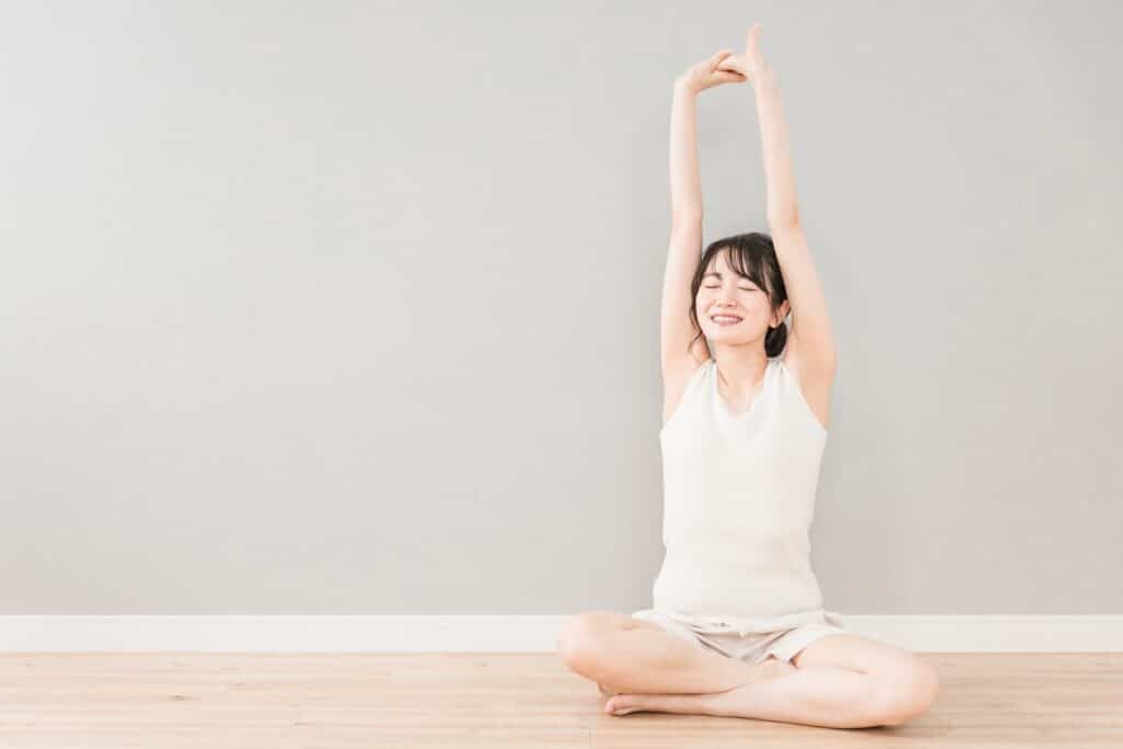 Woman sitting cross-legged on the floor, stretching her arms upward with a relaxed smile, enjoying a refreshing morning stretch: PMR Relaxation