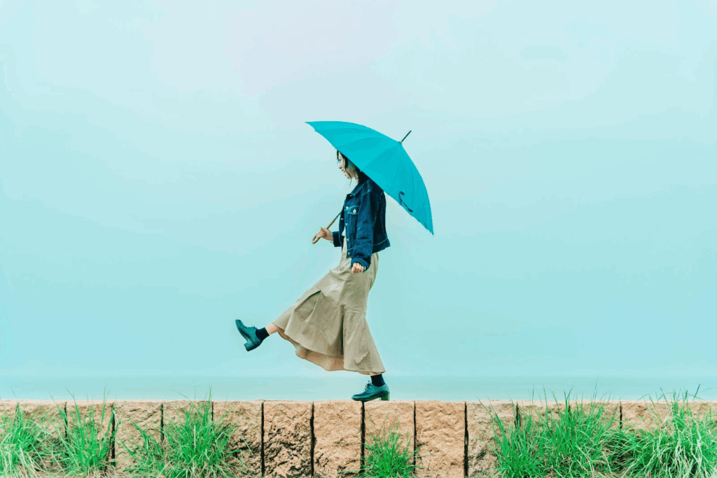 青い傘を差しながら歩道の縁を歩く女性。背景は淡い水色で、シンプルかつ幻想的な雰囲気。：マインドフルネス
