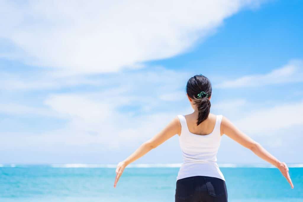Back view of a woman standing with arms outstretched by the sea, wearing a white tank top, relaxing and breathing deeply under the blue sky: PMR Relaxation