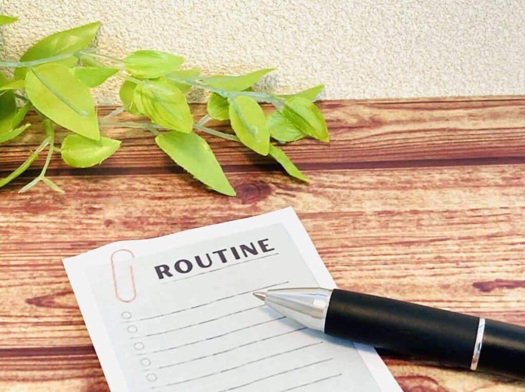 A black pen and a note paper titled "ROUTINE" are placed on a wooden desk, with a green decorative plant beside them: PMR Relaxation
