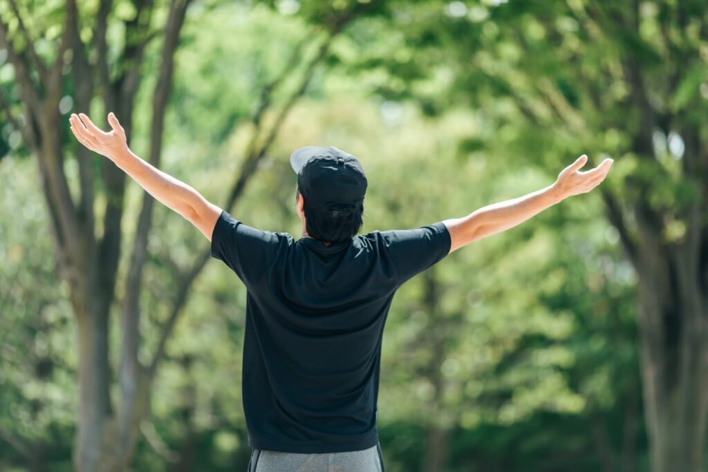 木々の緑に囲まれた屋外で、両腕を大きく広げて立つ男性の後ろ姿:不安を和らげる瞑想