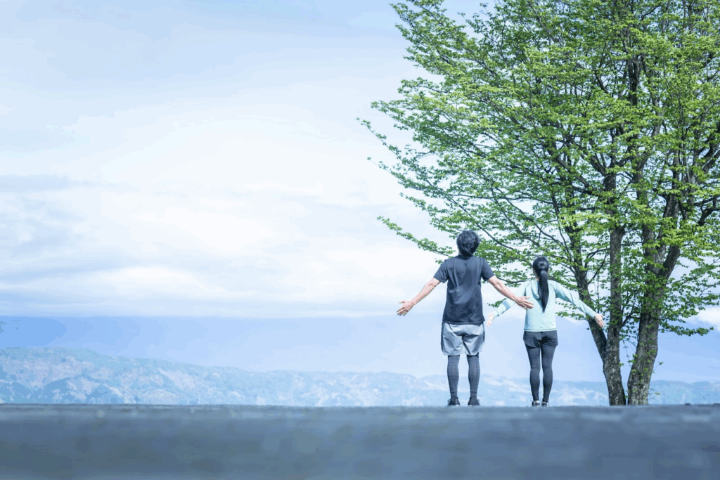 青空の下、大きな木のそばで並んで立ち、両腕を広げる二人の人物。遠くに山々が見える:セルフコンパッション