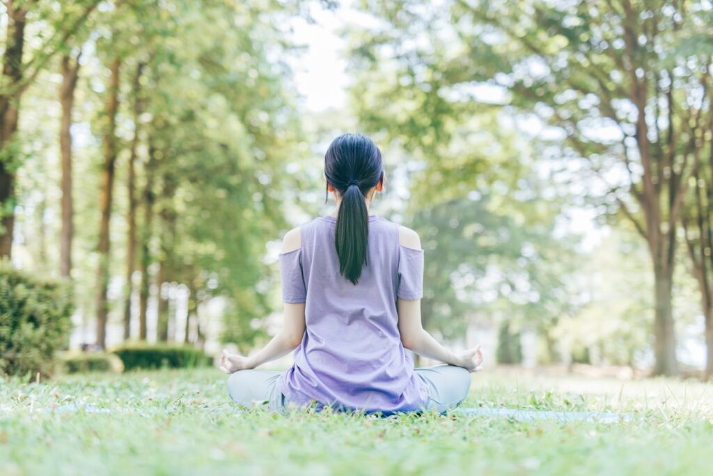 A woman in a purple outfit is sitting cross-legged on the grass, meditating in a park surrounded by trees: Joe Dispenza Meditation