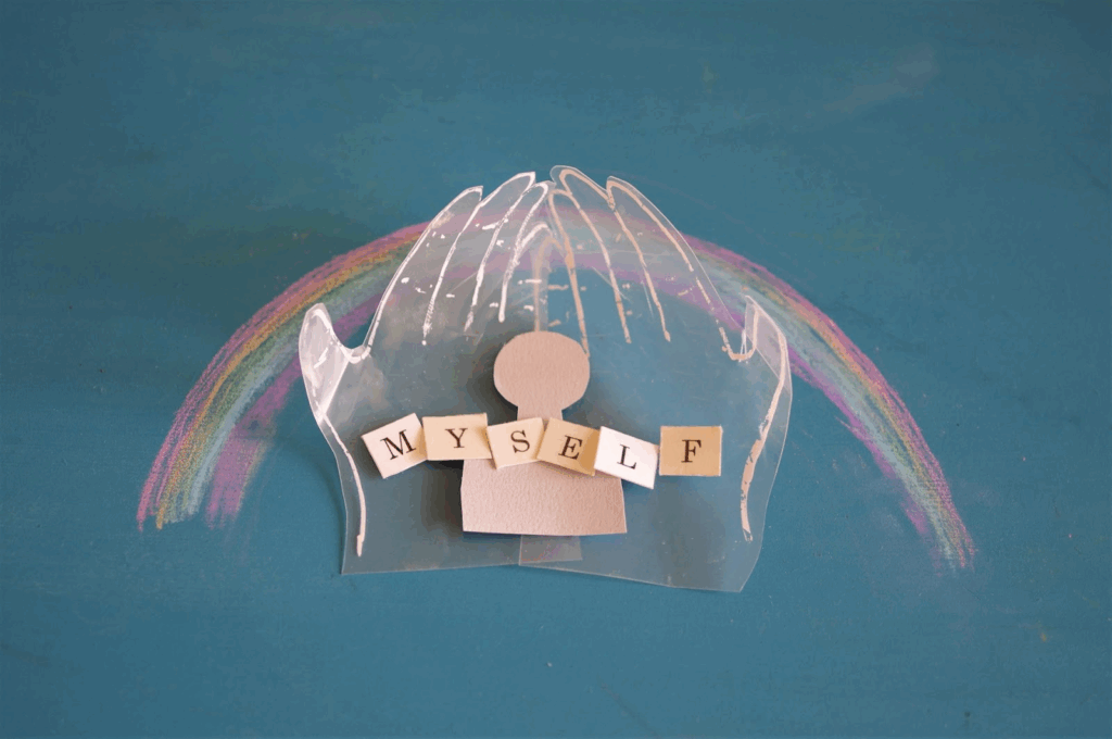 A paper cutout figure with the word "MYSELF" placed in front, protected by transparent illustrated hands, with a rainbow drawn on a blue background. :Tara Brach