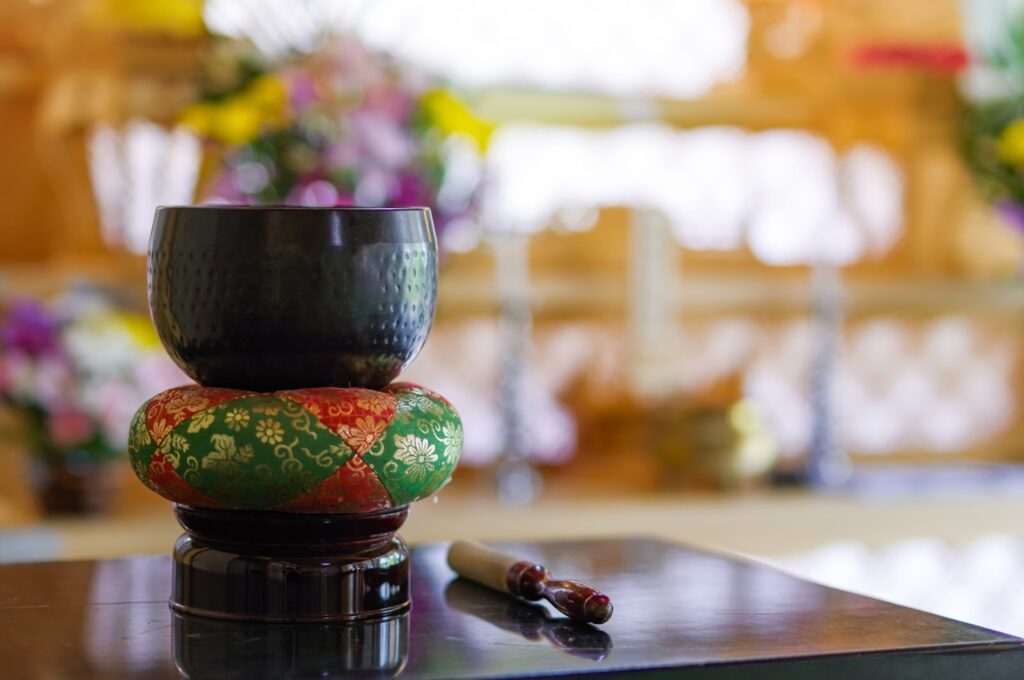 お寺で使われるおりんが、赤と緑の座布団の上に置かれている。背景にはぼんやりとした花が見える:睡眠のための穏やかな音楽