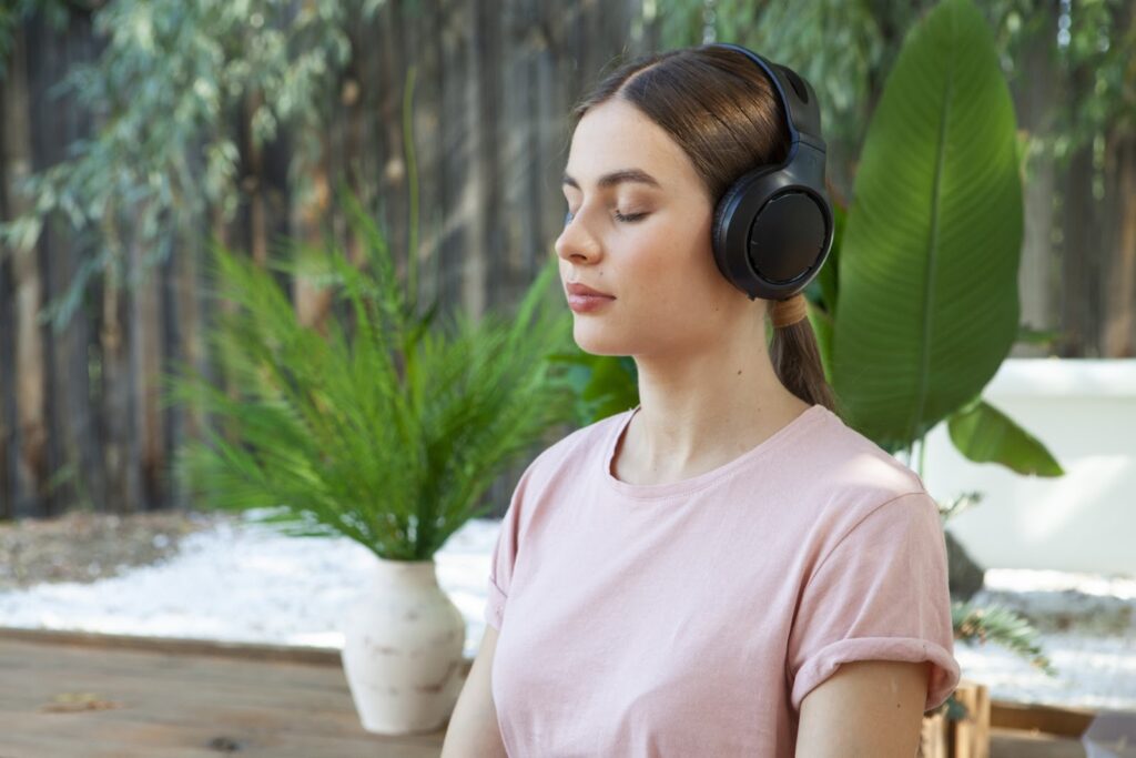 A woman wearing headphones with her eyes closed, meditating and relaxing outdoors, with potted plants in the background: Peace and Mind