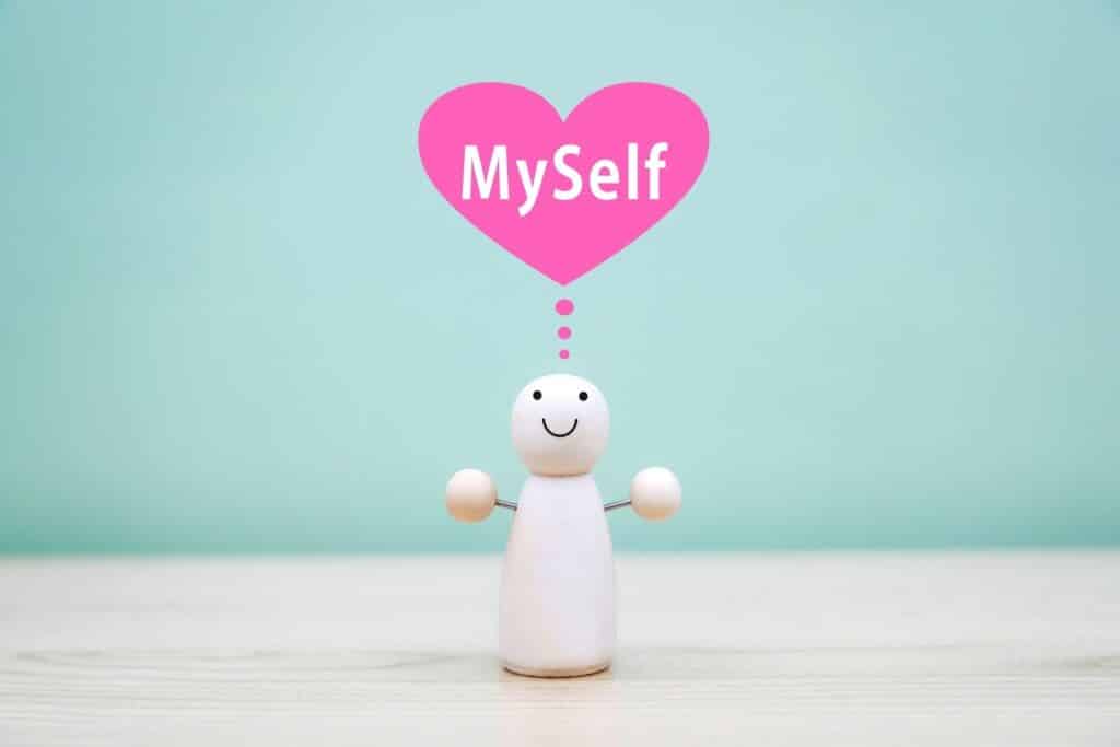 木製の小さな人形が笑顔で立ち、頭上には「MySelf」と書かれたピンクのハートが浮かんでいる。自己愛やセルフケアの象徴的なイメージ:心の平安