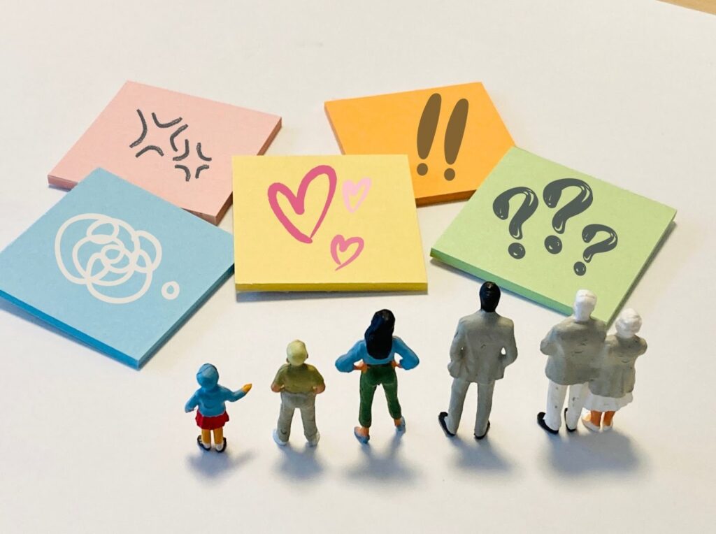 色とりどりの付箋に感情や記号(怒り、混乱、ハート、驚き、疑問)が描かれ、前に並んだ小さな人形たちがそれを見上げている様子。感情や思考の多様性を表現:心の平安