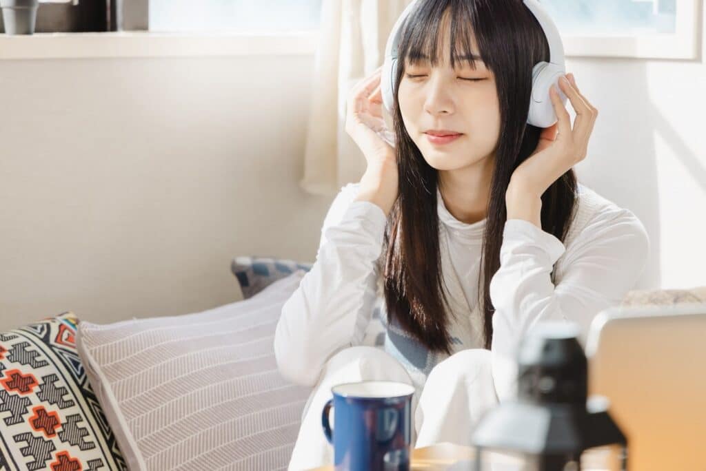 白いヘッドホンをつけて目を閉じ、リラックスして音楽を聴いている女性。日差しが差し込む部屋で穏やかな表情を浮かべている:心の平安