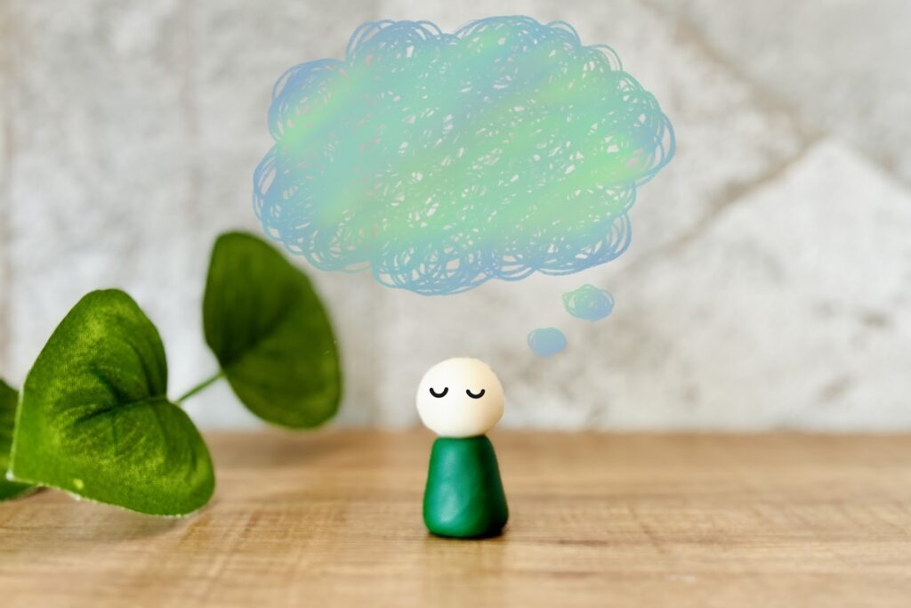 A small figurine with closed eyes sits on a wooden table, with a blue and green cloud-like illustration above its head representing thoughts. A green leaf is visible on the left side: Joe Dispenza Meditation