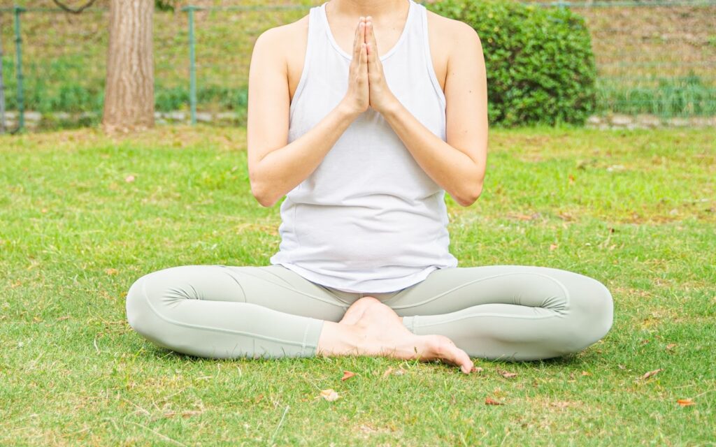 A woman sitting cross-legged on the grass, meditating with her hands pressed together in front of her chest. She is wearing a white tank top and light green leggings: Joe Dispenza Meditation