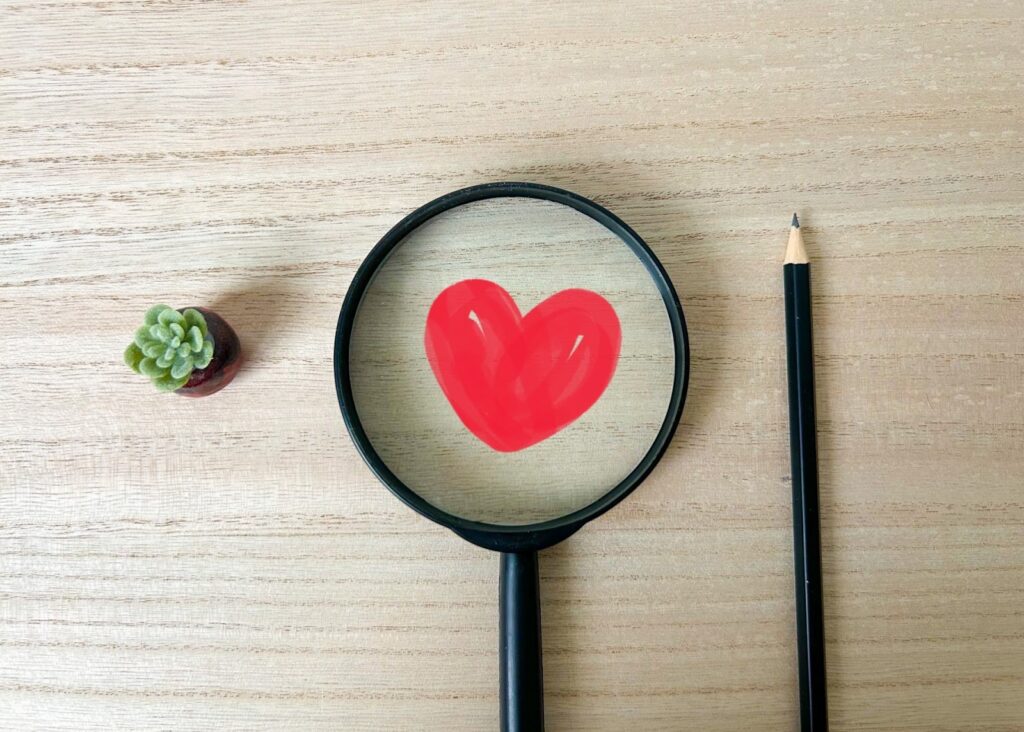 A magnifying glass on a wooden desk enlarging a red heart. Next to it are a pencil and a small potted succulent plant: Joe Dispenza Meditation