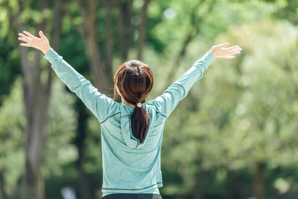 A woman with a ponytail stands in a lush green park, raising her arms wide and looking up at the sky, seen from behind : Breathing Techniques for Anxiety