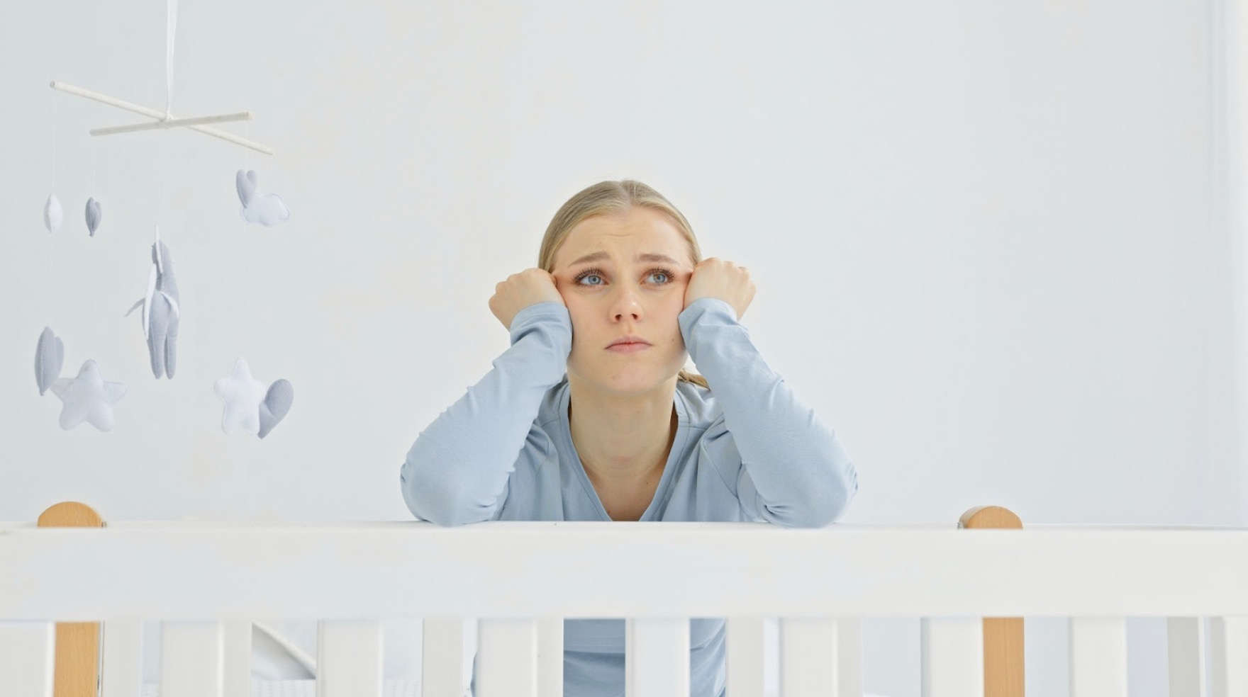 A young mother rests her face in her hands, looking worried while leaning on a white baby crib. The background is light blue and white, with a mobile hanging above the crib. :Anxiety Disorder Meditation