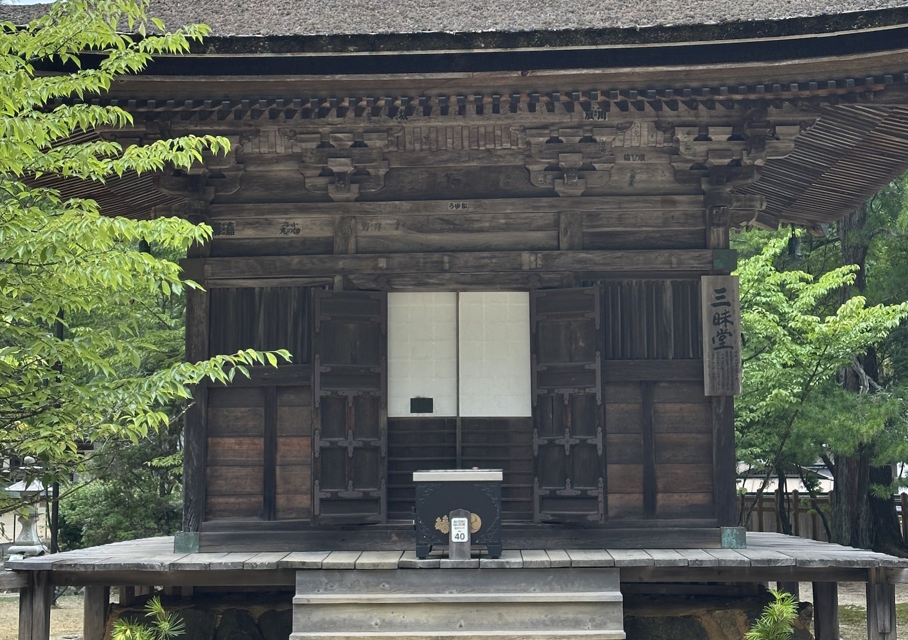 An old wooden temple building with a thatched roof, surrounded by tall trees, with stone steps leading up to the entrance.