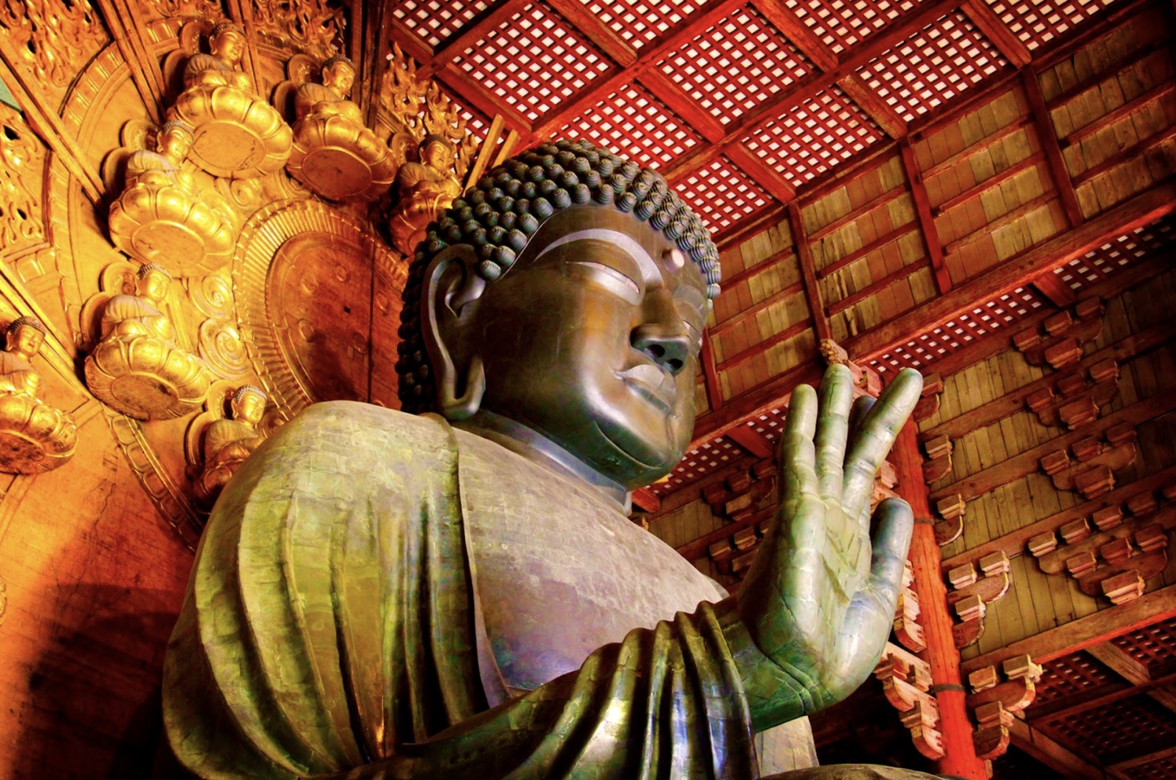The Great Buddha (Vairocana Buddha) of Tōdai-ji in Nara, Japan, raising its right hand in the Abhaya mudra. Behind the statue, golden Buddha figures and intricate decorations create a majestic and sacred atmosphere. :Japanese Meditation