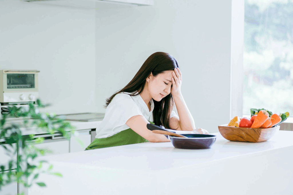A woman wearing an apron sits in the kitchen with her hand on her forehead, looking tired. In front of her are a frying pan and a wooden bowl filled with vegetables.: Subconscious