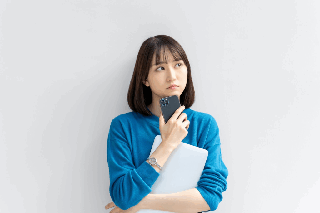 A woman in a blue sweater holding a laptop and pressing a smartphone to her chin, appearing thoughtful. She is standing against a plain white background. :Meditation Timer 
