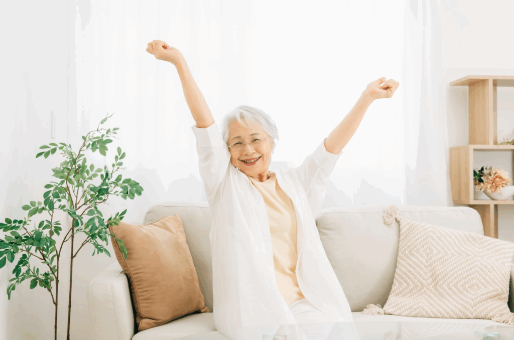 ソファに座り、両腕を伸ばして笑顔でストレッチする高齢の女性：深呼吸