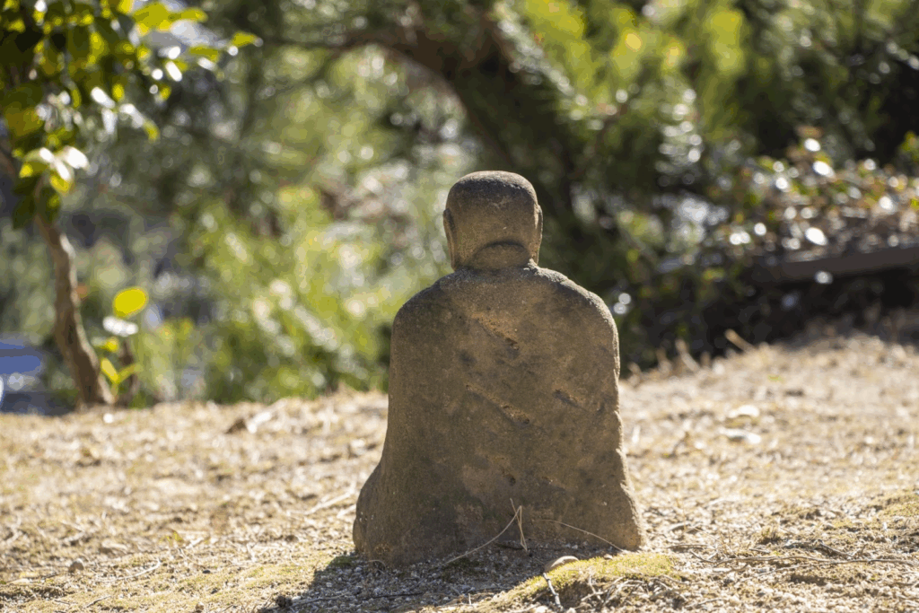 苔むした小さな石仏が、背中を見せて座っている。背後には柔らかな木々の緑が広がり、静かな雰囲気が漂っている。：日本の瞑想
