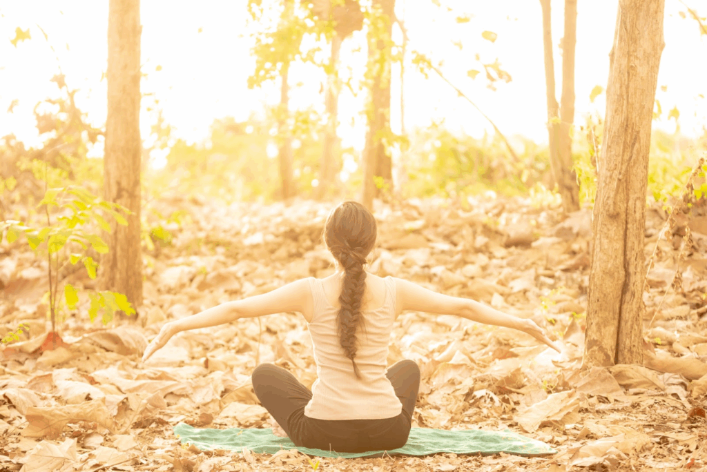 A woman sits on a yoga mat in a forest covered with autumn leaves, stretching her arms wide as she faces the morning sunlight. The scene glows in warm golden tones. :Meditation for Stress and Anxiety