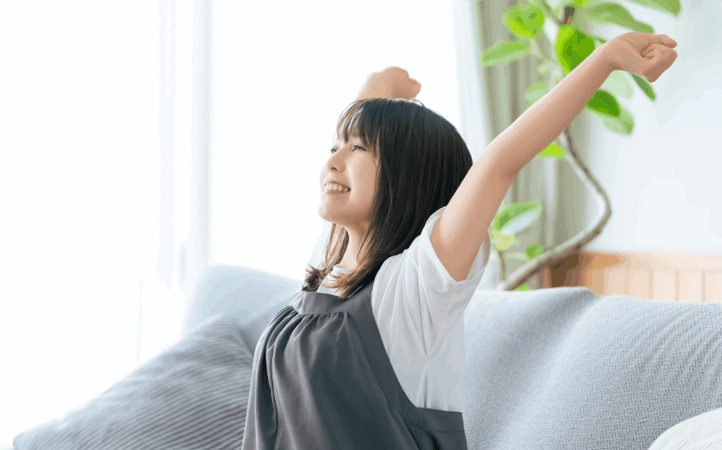 A young woman sitting on a sofa, stretching with a big smile. She looks relaxed in a bright room, expressing a refreshing morning mood. :Meditation Retreat