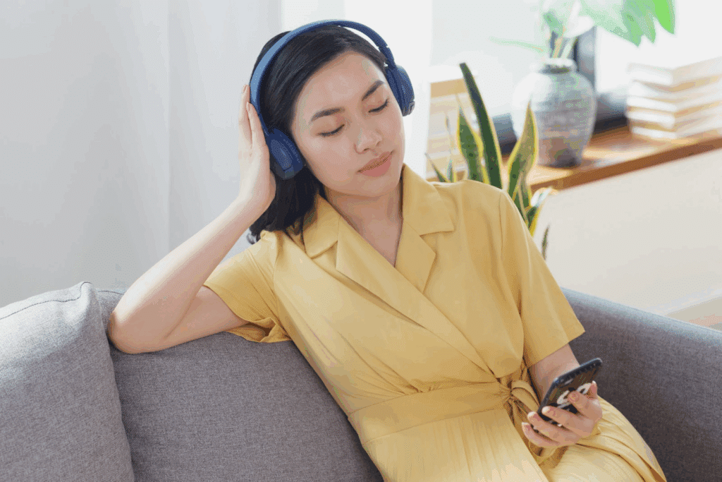 A woman in a yellow dress sitting on a sofa, relaxing with her eyes closed while listening to music. She is wearing blue headphones and holding a smartphone in her hand. :Meditation Retreat