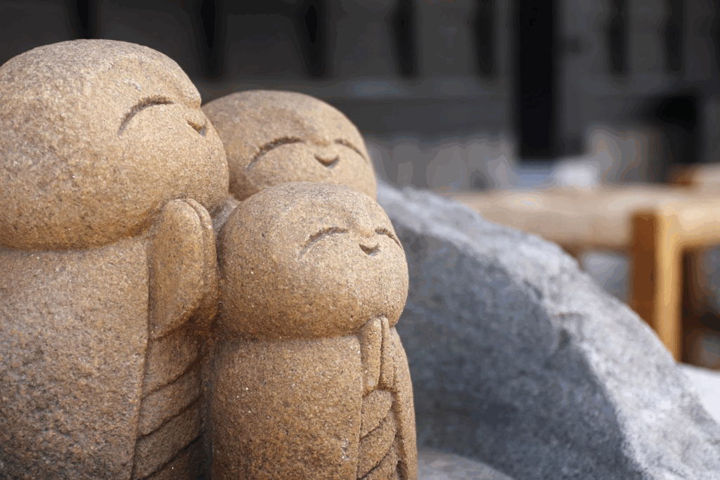 Three small stone statues of Jizo with gentle smiling faces. They hold their hands together as if in prayer, radiating a sense of peace and comfort. :Meditation Retreat