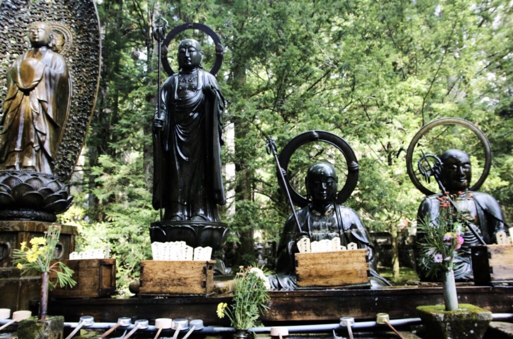 木々に囲まれた場所に並ぶ複数の仏像。手前には花や柄杓が供えられている。：日本の瞑想