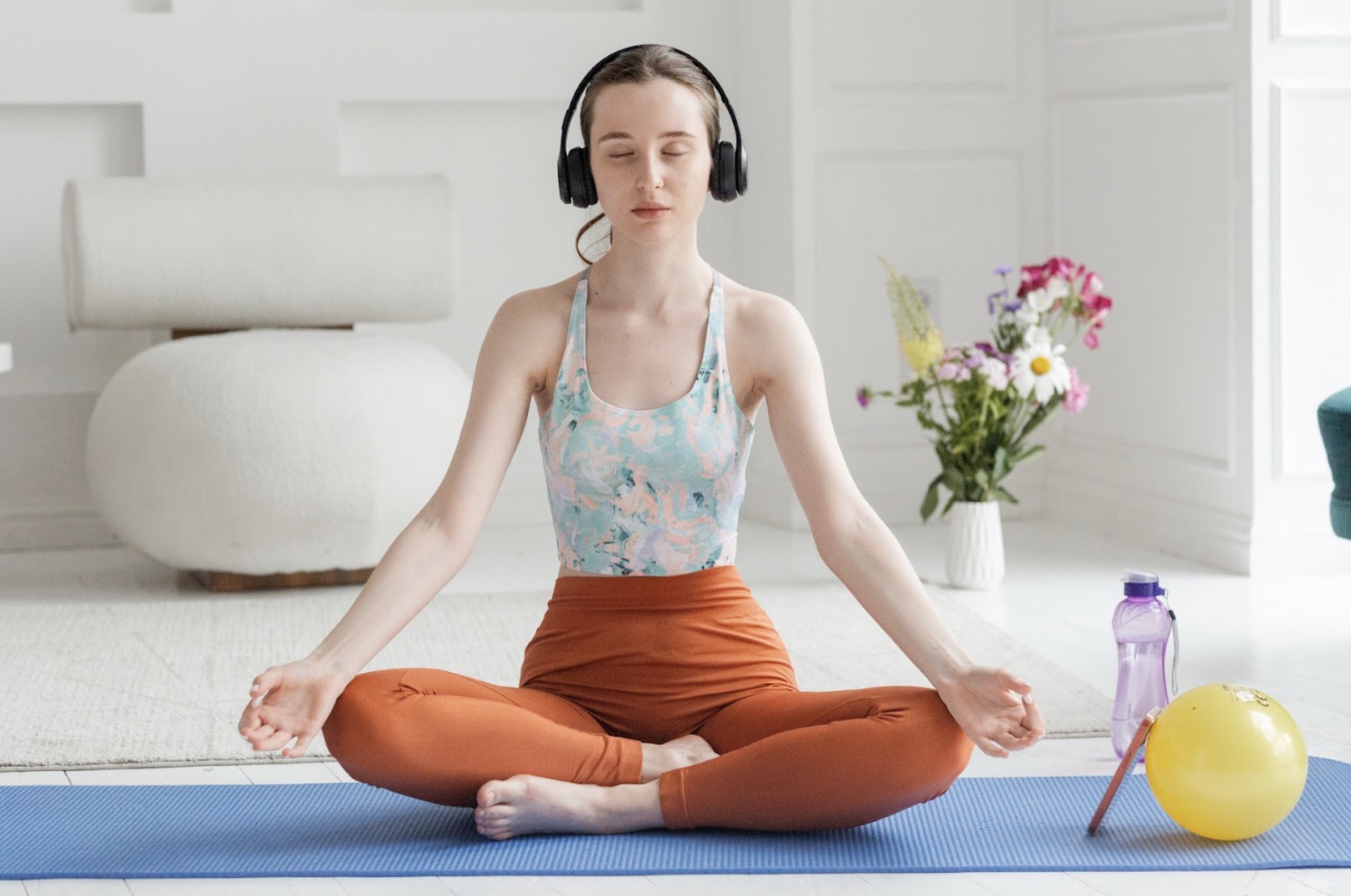 A woman sitting on a yoga mat, meditating with her eyes closed while wearing headphones. Nearby are a vase of flowers, a water bottle, and a yellow ball. :Guided Meditation
