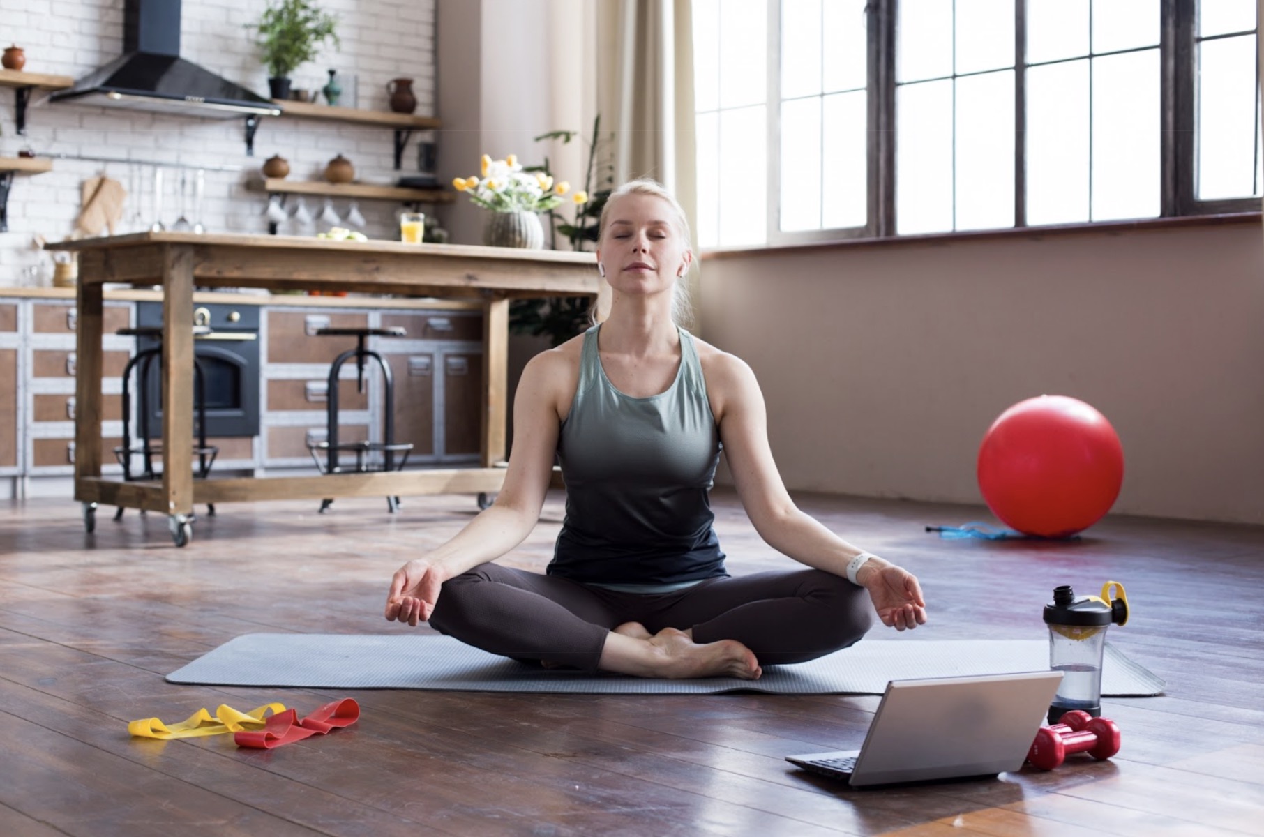A woman sitting cross-legged on a yoga mat in a living room, meditating with eyes closed, next to a laptop and fitness equipment.