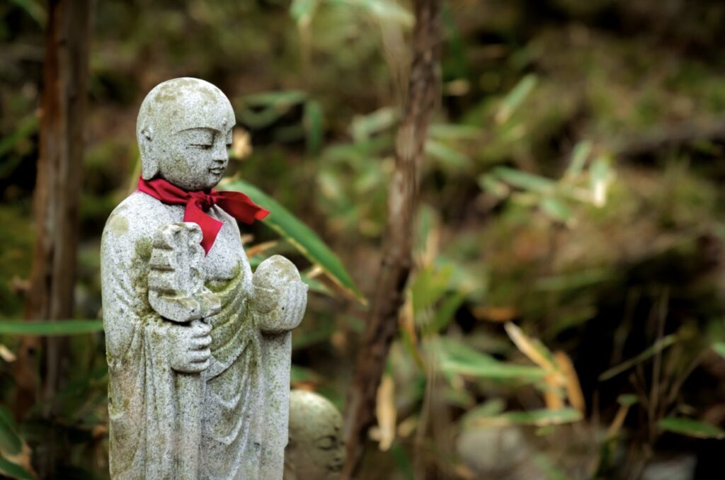 A moss-covered stone statue of Jizo Bodhisattva, wearing a red cloth around its neck and holding a staff and a wish-fulfilling jewel. The statue stands quietly in a natural setting with bamboo and greenery in the background.:Subconscious