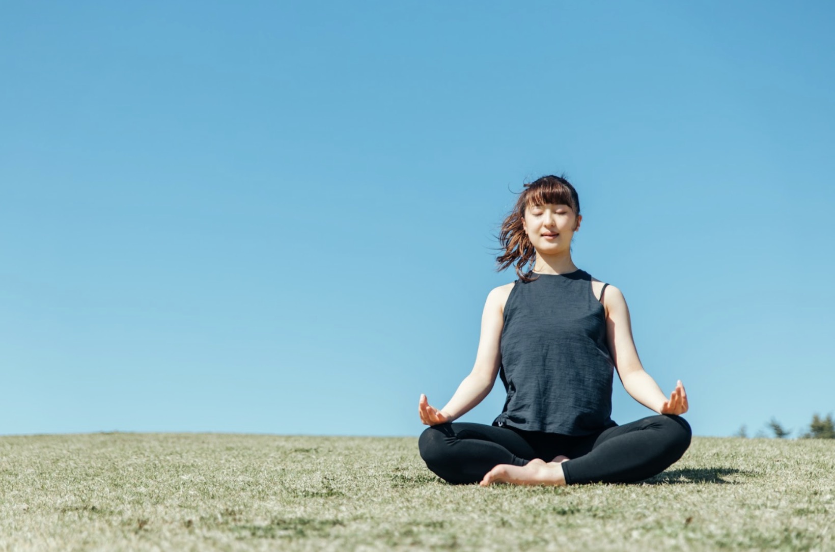 A woman sitting cross-legged on grass in a meditation pose, eyes closed, under a clear blue sky. :Diaphragmatic Breathing