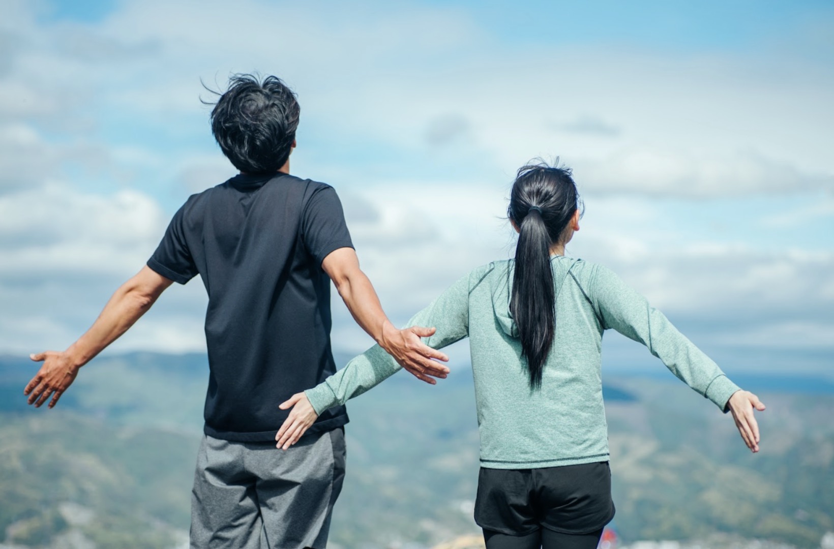 山の景色を背に並んで立ち、両腕を広げる男女の後ろ姿：深呼吸
