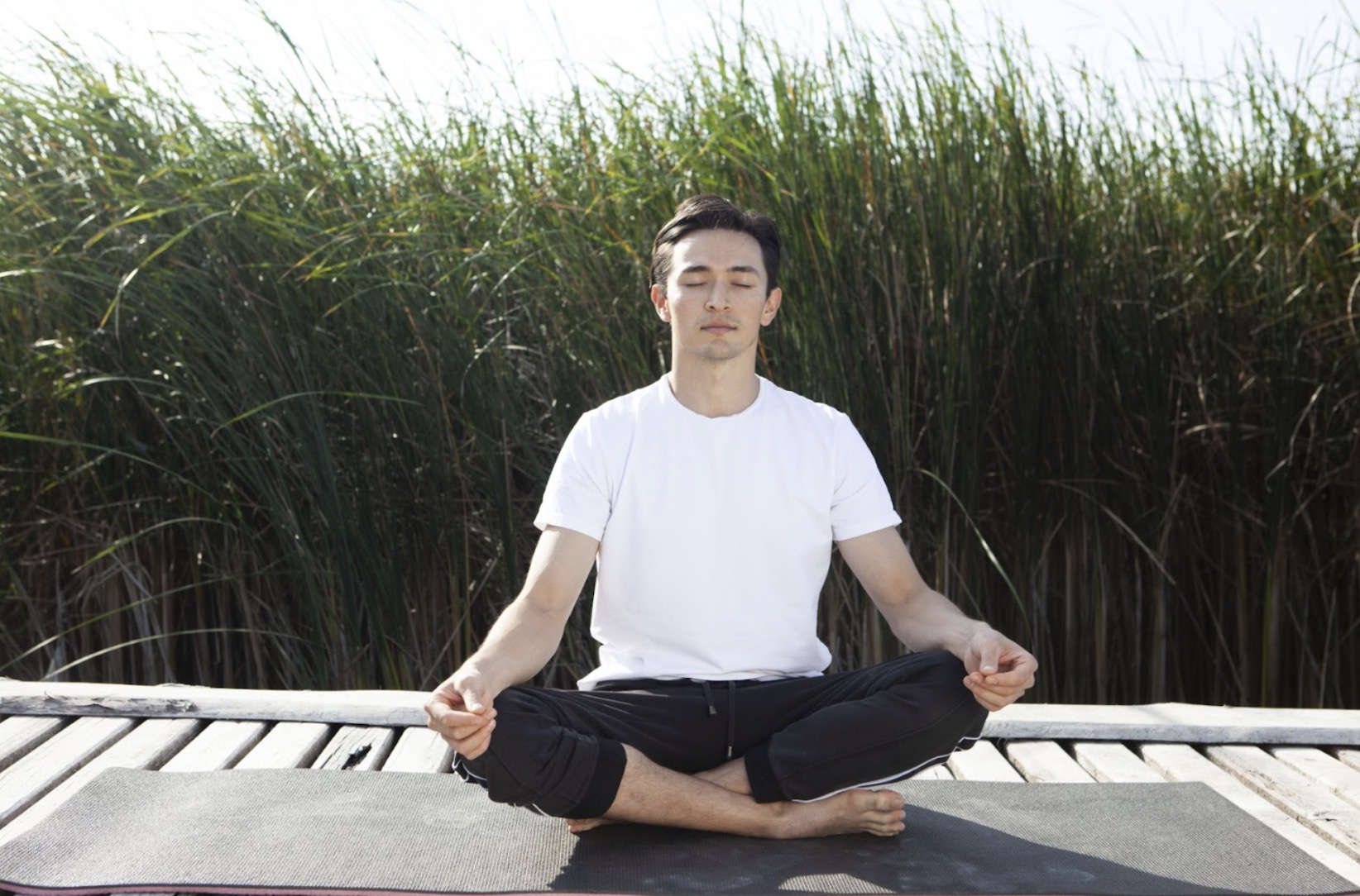 A man in a white T-shirt sits cross-legged on a yoga mat outdoors, meditating with closed eyes, surrounded by tall grass in the background : Body Scan Meditation
