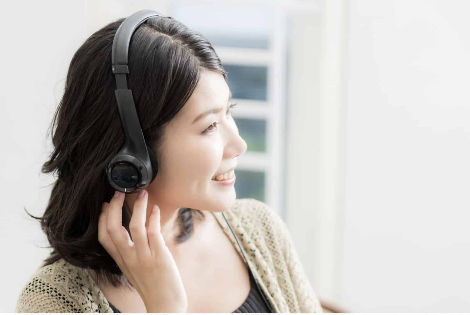 ヘッドフォンをつけて音楽を聴きながら微笑む女性。明るい自然光が差し込む室内で、リラックスした表情を見せている：ガイド付き筋弛緩法