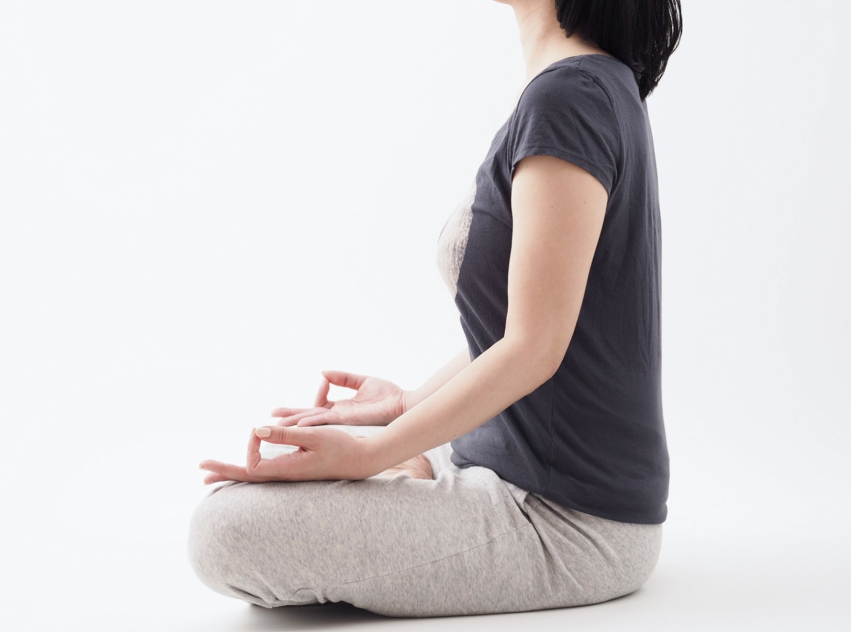 A woman wearing gray pants and a black T-shirt sits cross-legged against a white background, maintaining an upright posture with her hands resting on her knees in a meditative gesture. :Meditation Positions