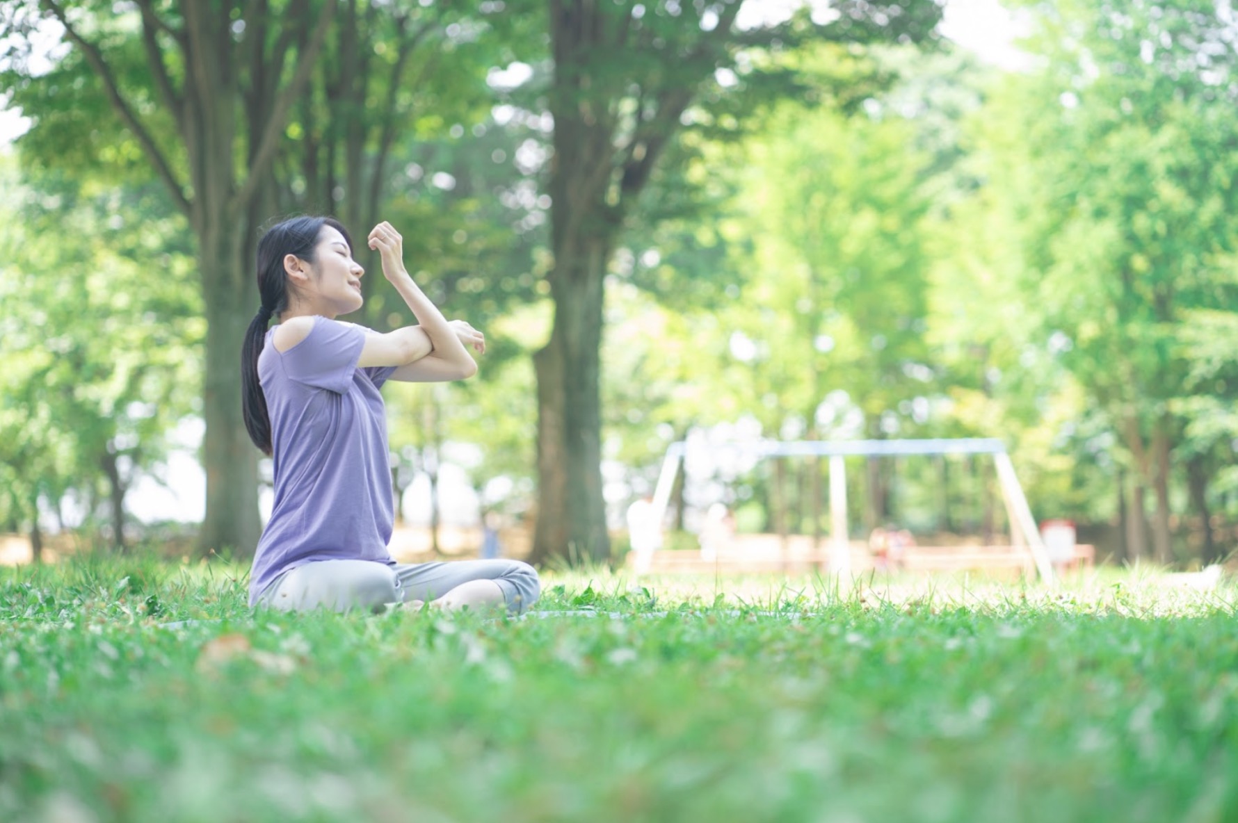 A young woman sitting on the grass in a park, stretching her arms, surrounded by lush green trees : Mindfulness