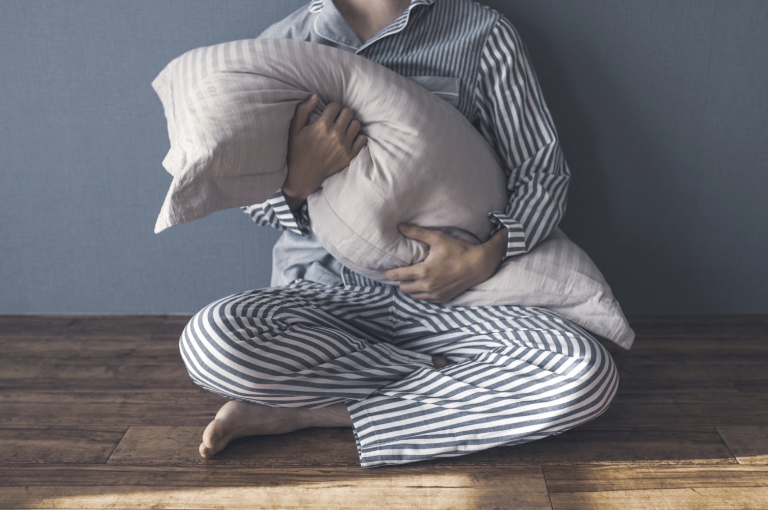 ボーダー柄のパジャマを着た人物が床に座り、抱きしめるように枕を持っている：睡眠催眠