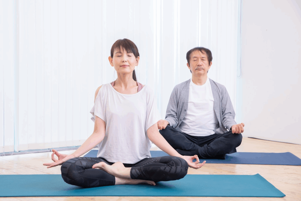 A middle-aged man and woman sit cross-legged on yoga mats in a bright room, eyes closed in meditation. Their calm expressions convey a sense of inner peace and mindful breathing. :Meditation Positions