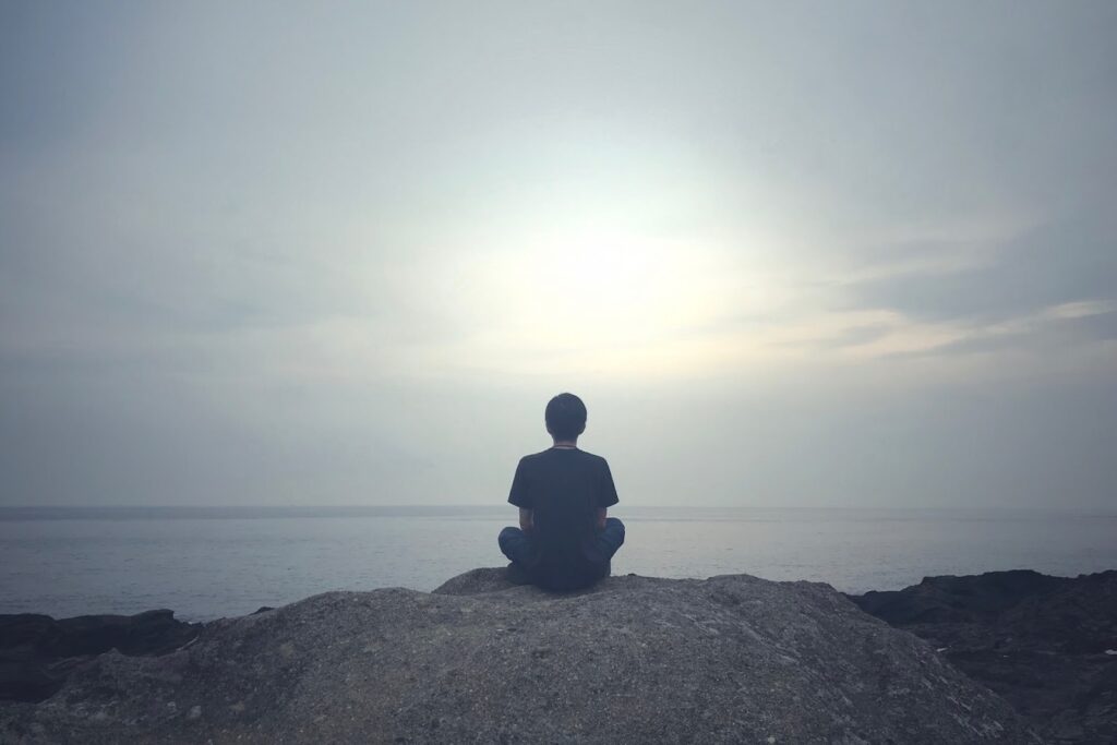 A person sitting on a rock by the sea, meditating while gazing at the horizon under a pale gray sky, evoking a sense of calm and stillness :Meditation for Anxiety