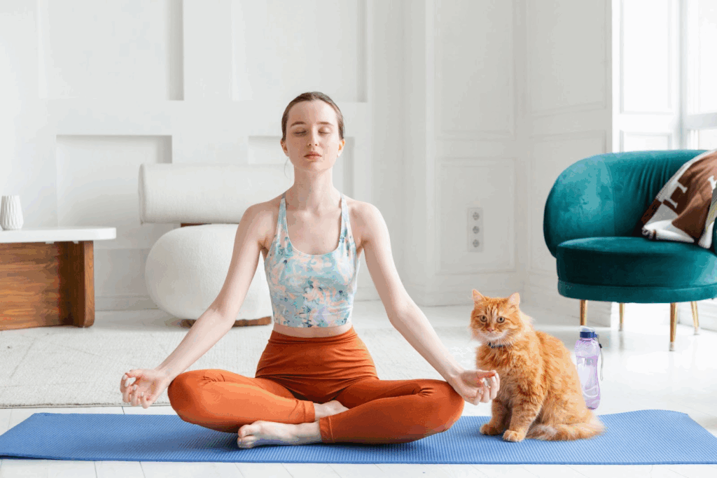 A woman sits cross-legged on a yoga mat with her eyes closed in meditation, while an orange cat sits calmly beside her. The bright room radiates a sense of morning tranquility and relaxation :Meditation Positions