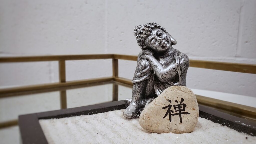 A small Buddha statue sitting peacefully beside a stone engraved with the word "Zen," placed in a miniature Zen garden with white sand : Mindfulness What Is It