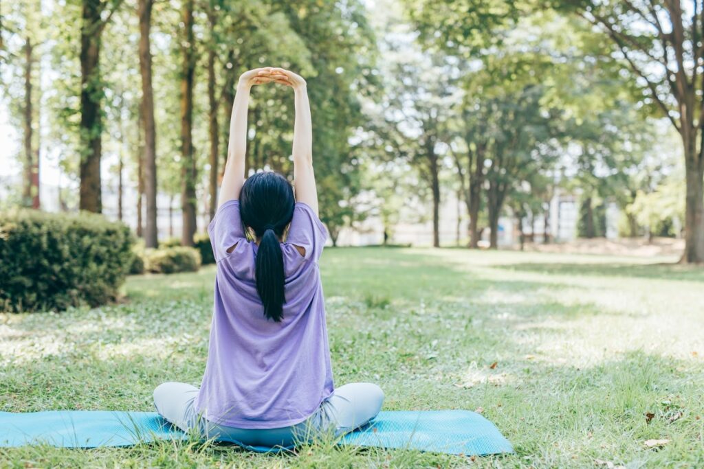A woman sitting on a yoga mat in a park, stretching her arms upward and taking a deep breath, surrounded by green trees in a peaceful natural setting : Mindfulness What Is It
