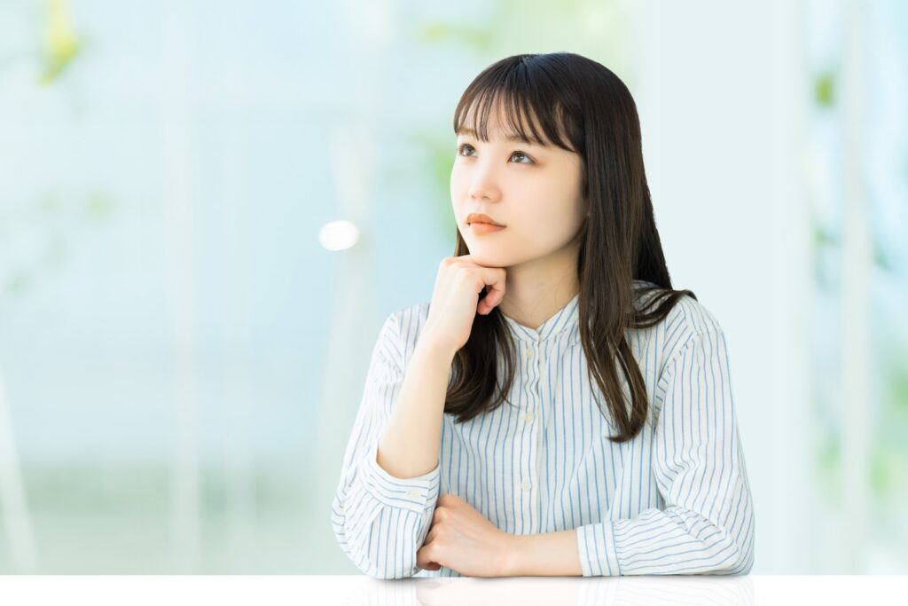 A young woman resting her chin on her hand, looking thoughtful as she gazes out in a bright indoor setting : Mindfulness What Is It