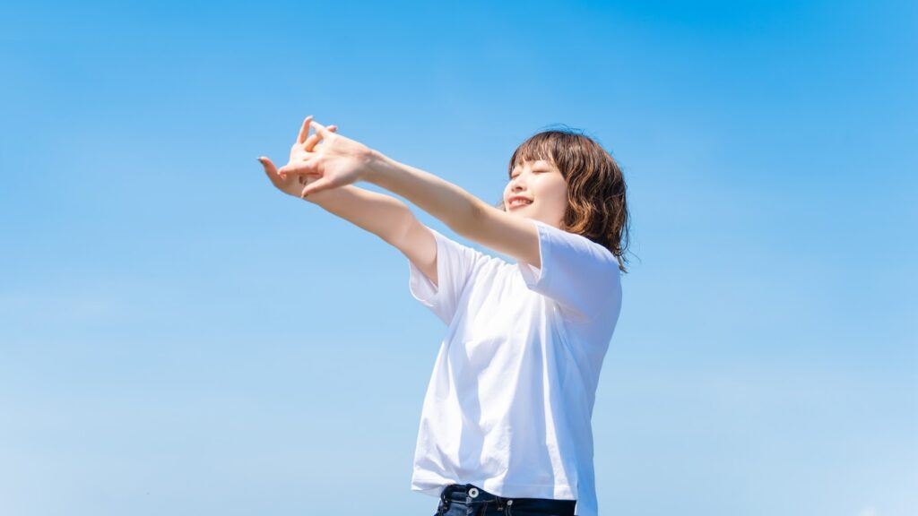 A woman in a white T-shirt stretching comfortably under a clear blue sky : How to Relax Anywhere