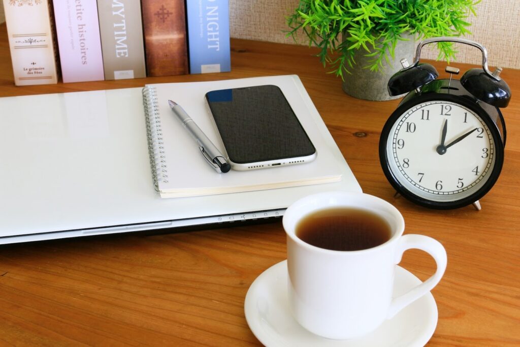 A cup of coffee on a wooden desk with a notebook, pen, smartphone, and a black alarm clock showing 12:10 p.m, representing a morning routine or a moment before starting work : How to Relax Anywhere