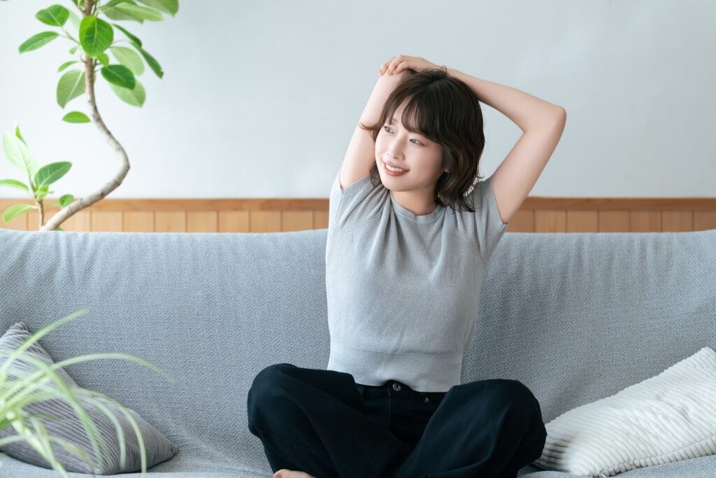 A woman in a gray top sits on a sofa, stretching her arms upward with a relaxed smile while looking to the side : Body Scan Meditation