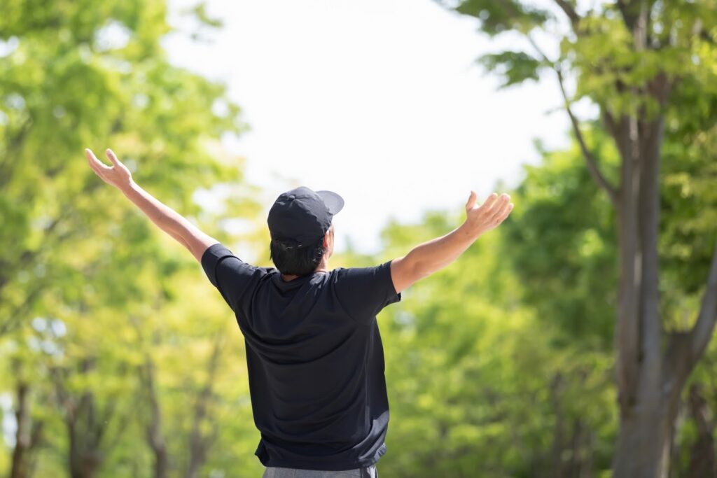 A man wearing a cap stands with his arms wide open, surrounded by trees in a park. His back is facing the camera, expressing a sense of freedom and connection with nature : Body Scan Meditation